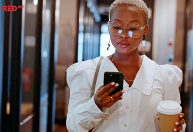 A stylish woman with short blonde hair, wearing glasses and a white blouse, walks through a modern office corridor while looking at her smartphone. She holds a takeaway coffee cup in her other hand. The “RED VIP” logo is positioned in the top-left corner.