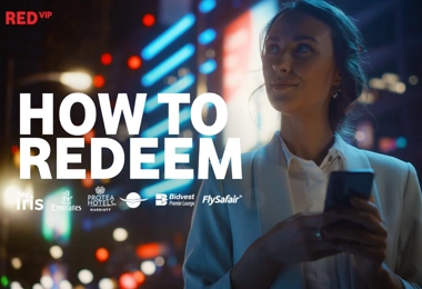 A young woman in business attire holds her smartphone while standing in a vibrant cityscape at night, illuminated by colorful neon lights. Bold white text overlay reads “HOW TO REDEEM,” with logos of various travel and hospitality brands, including Emirates, Protea Hotels, Bidvest Premier Lounge, FlySafair, and Iris, displayed beneath. The “RED VIP” logo is positioned in the top-left corner.